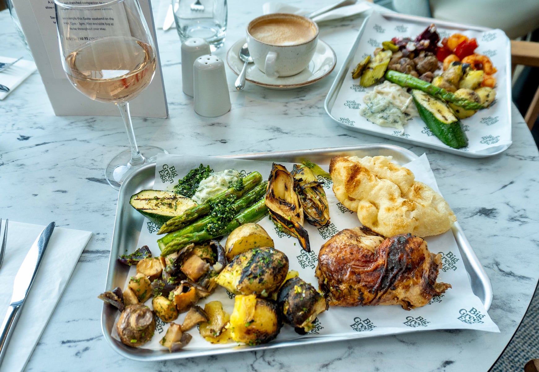 Grilled vegetables, roasted chicken, flatbread and potatoes, alongside a glass of rosé wine on a marble table.