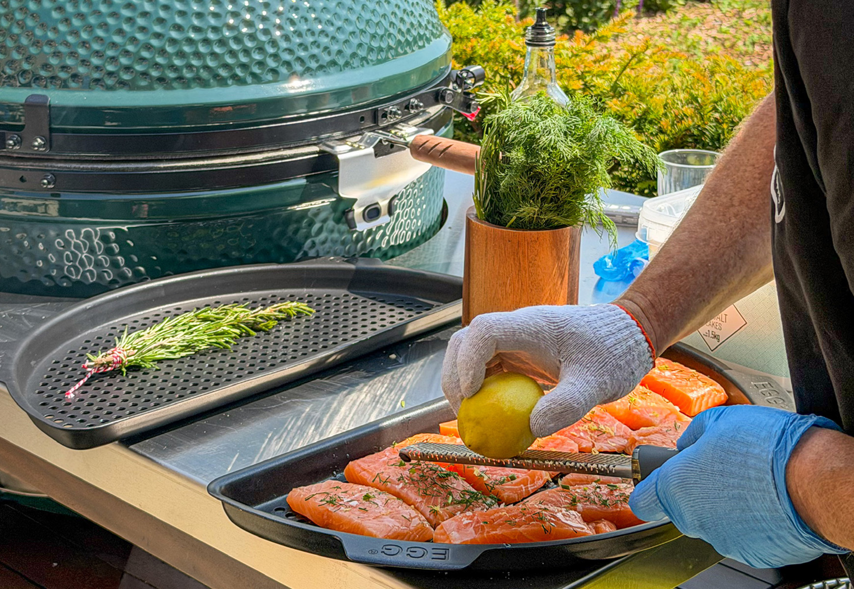 Chef preparing salmon fillets on a Big Green Egg station, zesting lemon over seasoned fish beside fresh herbs.