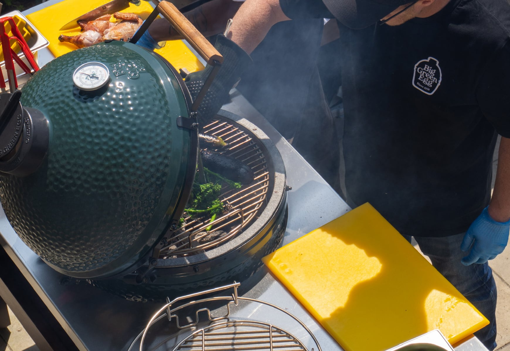 Open Big Green Egg barbecue grill with food cooking over coals, as a chef works at an outdoor cooking station.