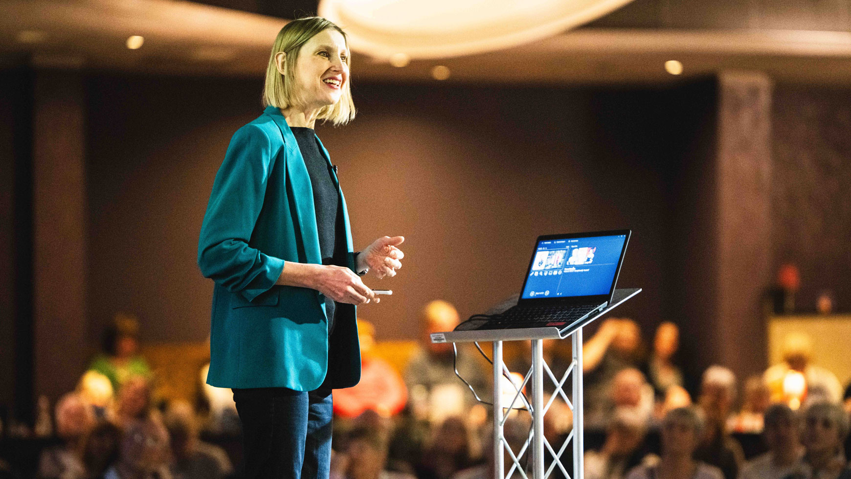 A presenter wearing a teal blazer stands on stage, gesturing toward a laptop while an audience watches attentively in the background