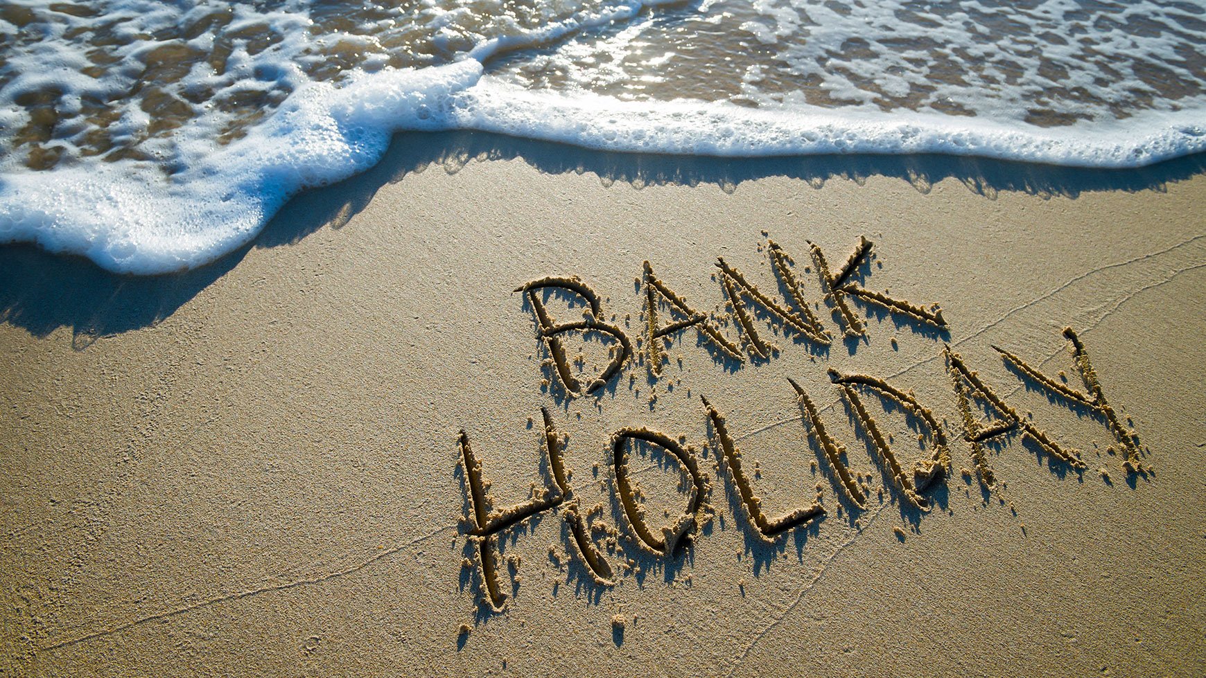 Bank holiday written in the sand with a wave approaching the message