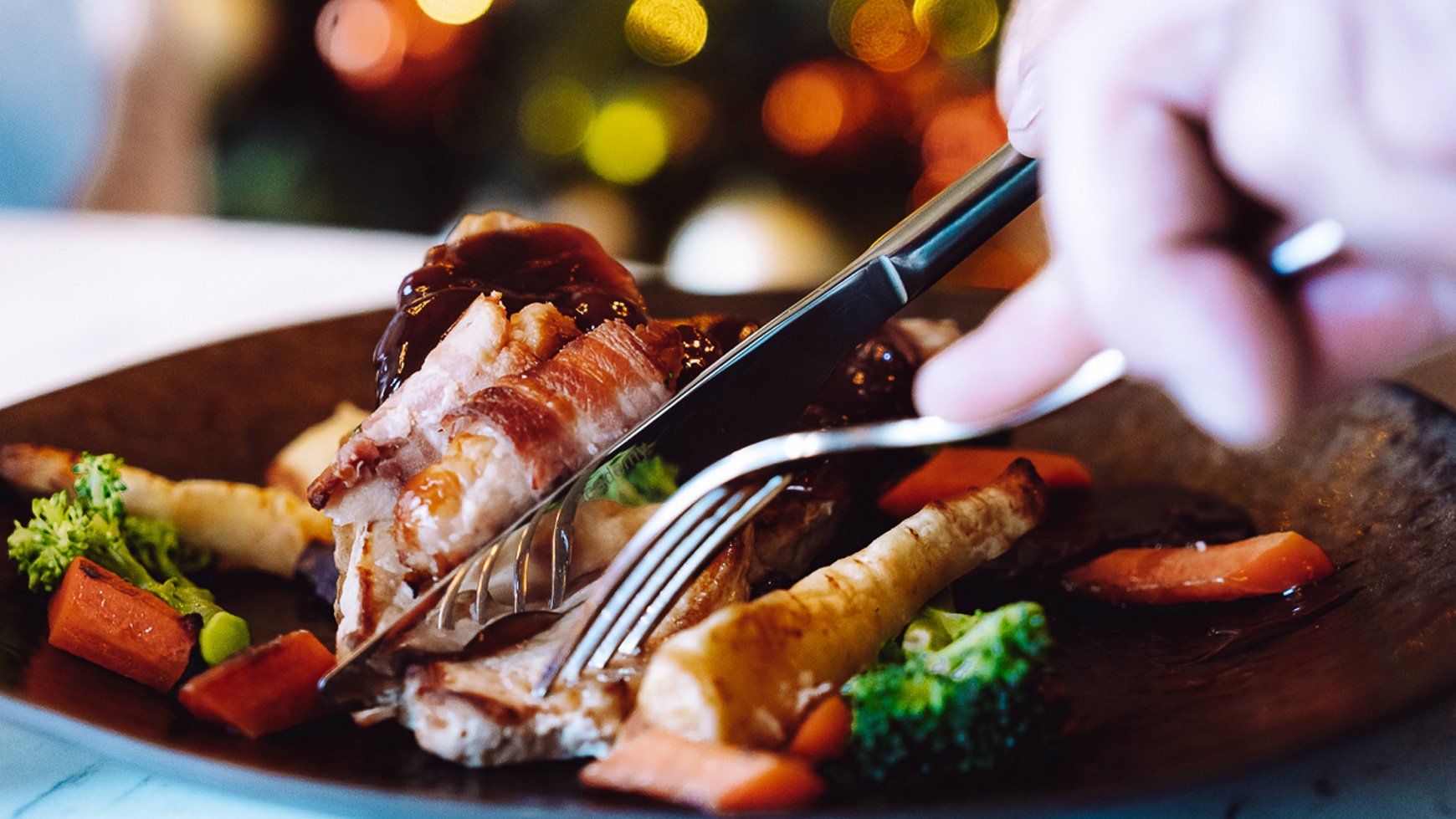 A hand using a knife and fork to cut into a plate of glazed meat with vegetables, set against a softly lit festive background.