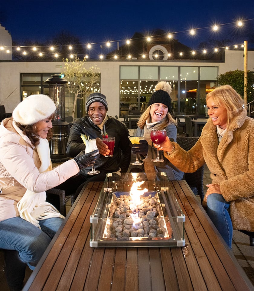 Four friends joyfully toasting drinks over a festive fire pit, creating a warm and inviting atmosphere.