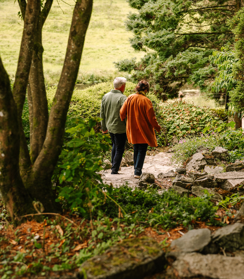A couple strolls hand-in-hand down a stone pathway, surrounded by lush greenery and trees.