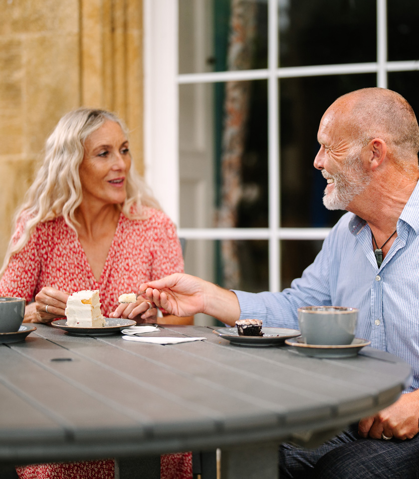 A couple shares dessert at an outdoor table, with coffee and cake on plates, in a cozy, inviting setting.