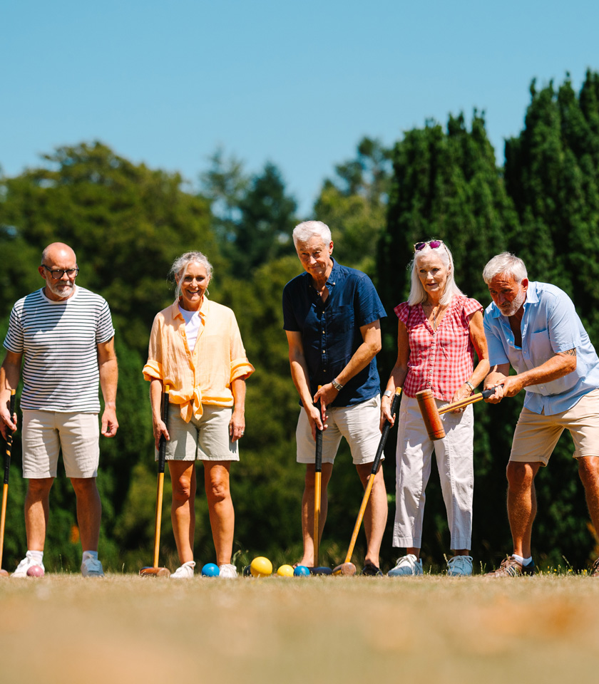 A group of five people stands on a sunny lawn, preparing to play croquet with colorful balls and mallets.