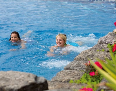 Two ladies swimming in a bright blue pool surrounded by lush greenery and vibrant flowers, creating a serene and relaxing atmosphere.