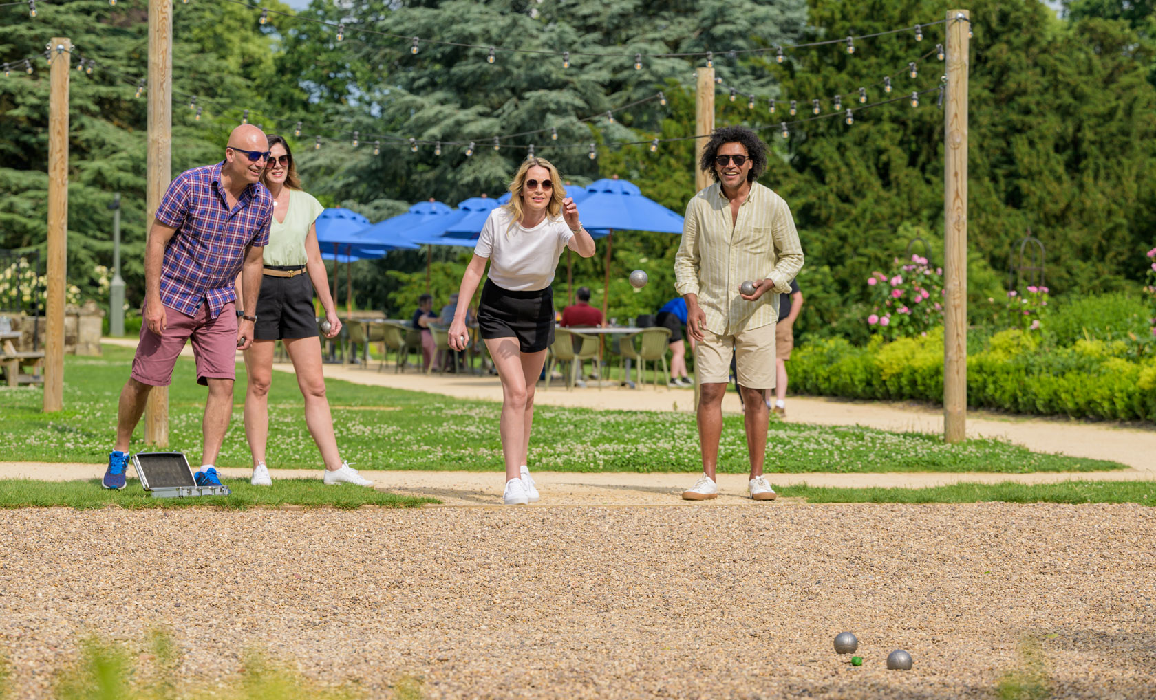 A group of four people plays pétanque on a sunny day at Studley Castle, with green trees and blue umbrellas in the background.