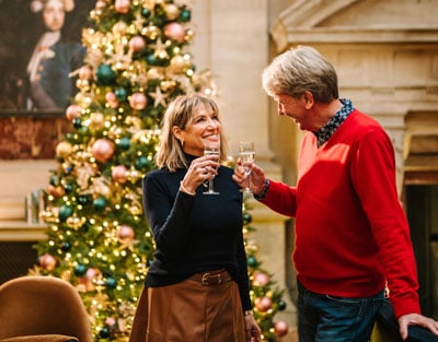 Four hands raise various cocktails in a toast, with a festive Christmas tree adorned with lights and ornaments in the background.