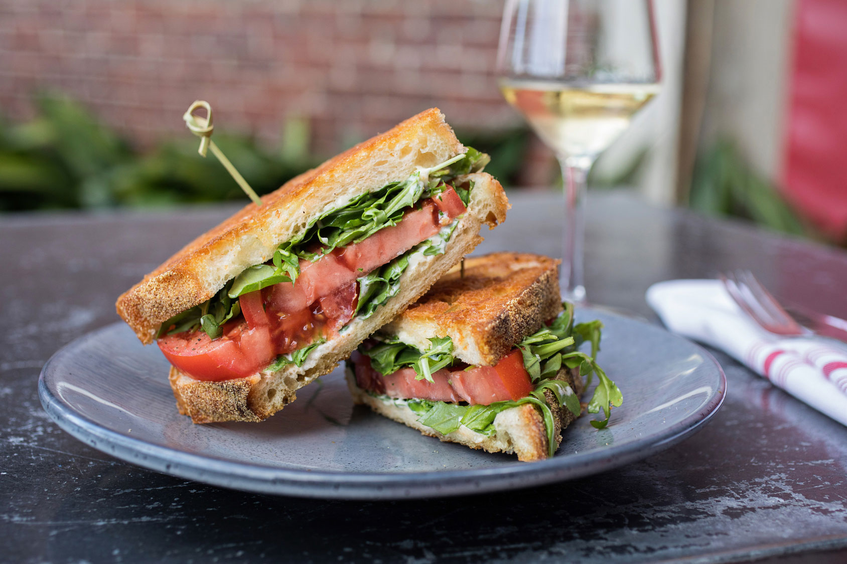 A close-up of a grilled sandwich with fresh tomatoes, greens, and a glass of white wine in the background, served on a gray plate.