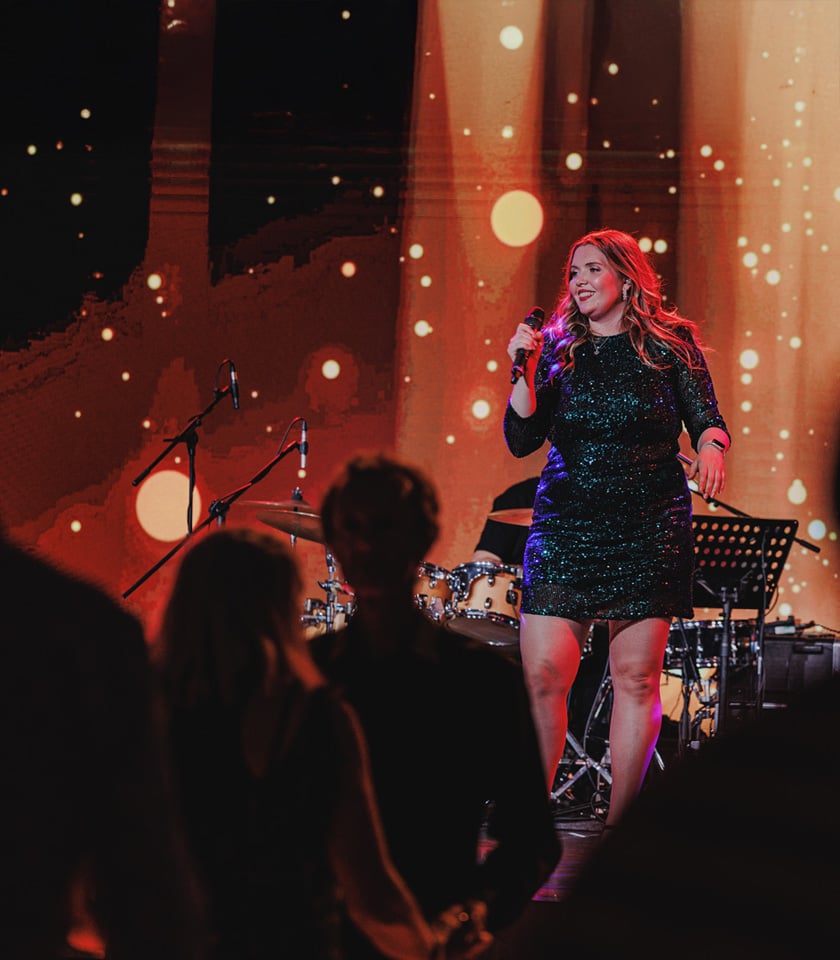 A woman in a sparkling green dress performs on stage with a microphone, surrounded by a colorful backdrop and musicians in the background.