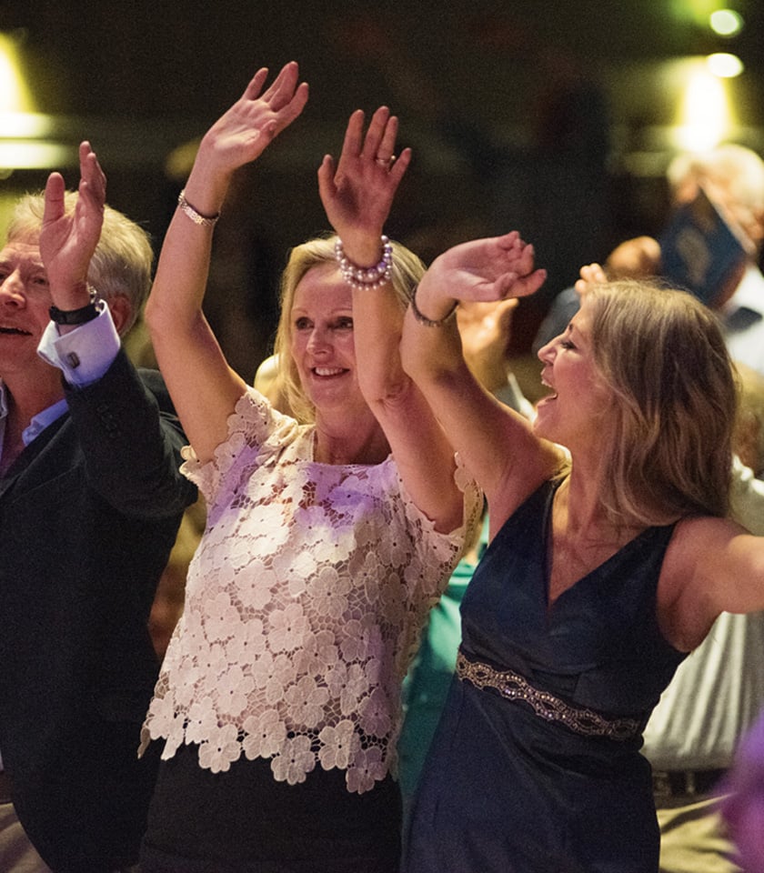A lively group of people joyfully dancing with raised hands, celebrating in a vibrant atmosphere. Colorful outfits and cheerful expressions are apparent.