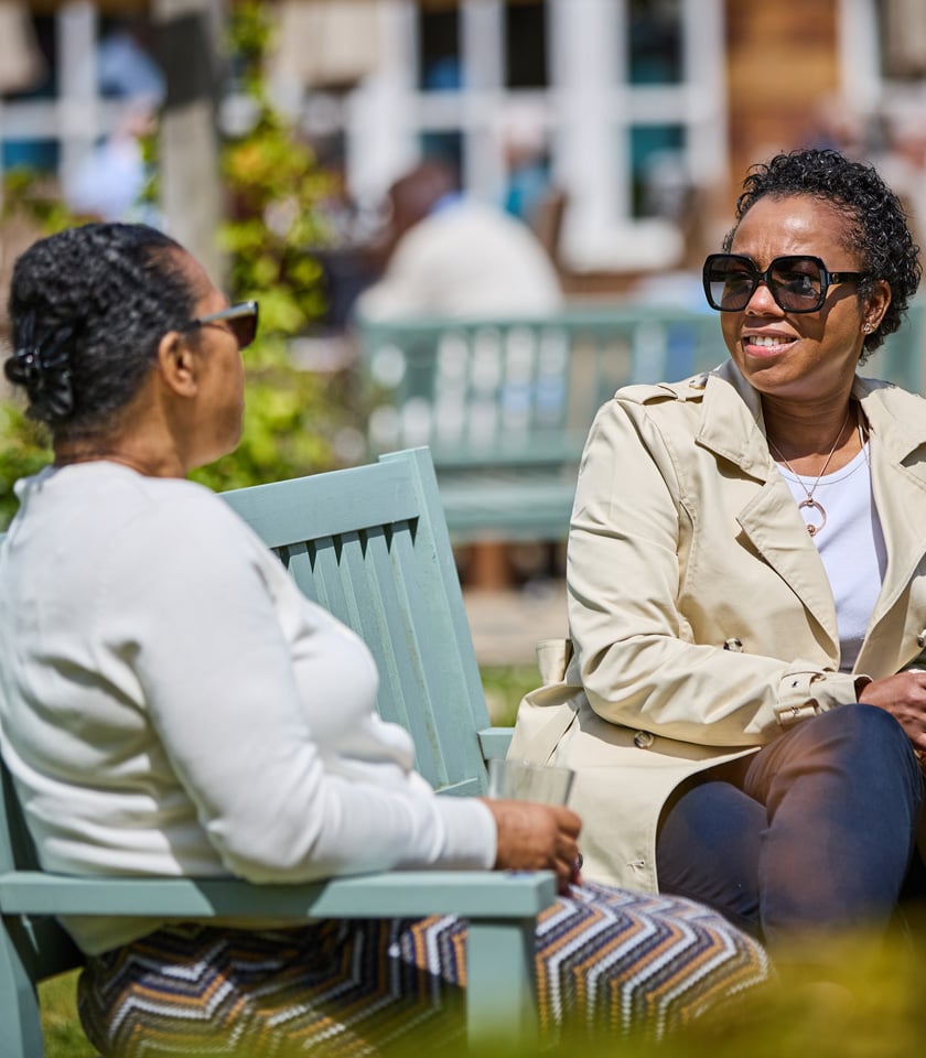 Two women sit outdoors on light blue benches, engaged in conversation under a sunny sky, surrounded by greenery and blurred background activity.