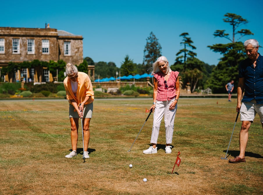 A group of four people plays pétanque on a sunny day at Studley Castle, with green trees and blue umbrellas in the background.