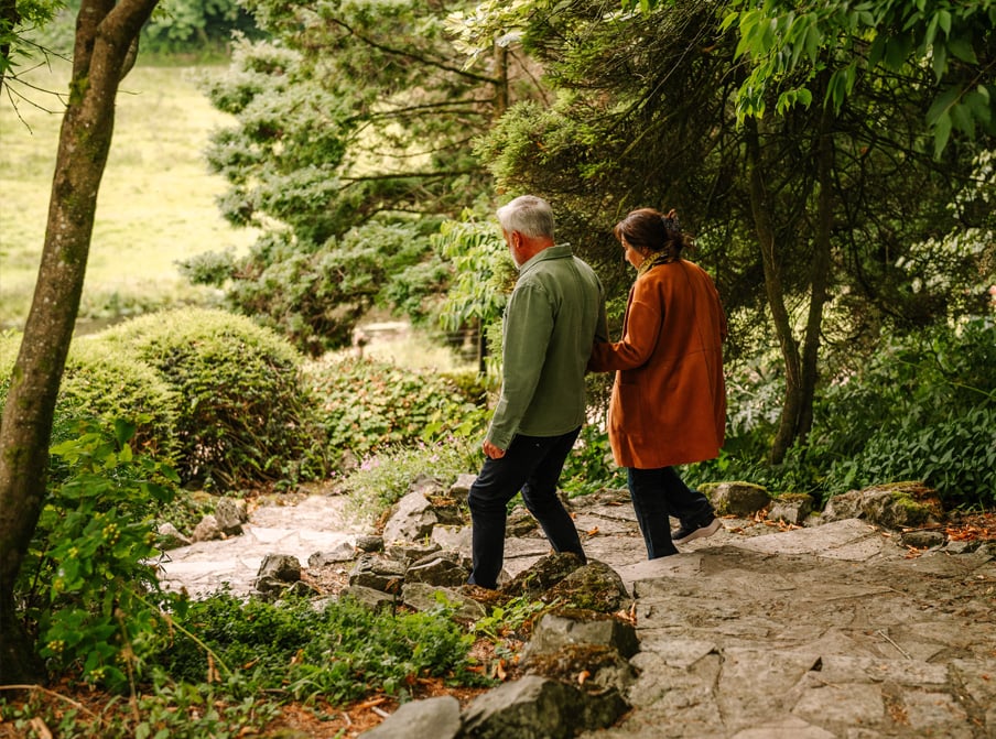 A man and a woman walk hand-in-hand down a stone path surrounded by lush greenery and ivy-covered bushes in a serene landscape.